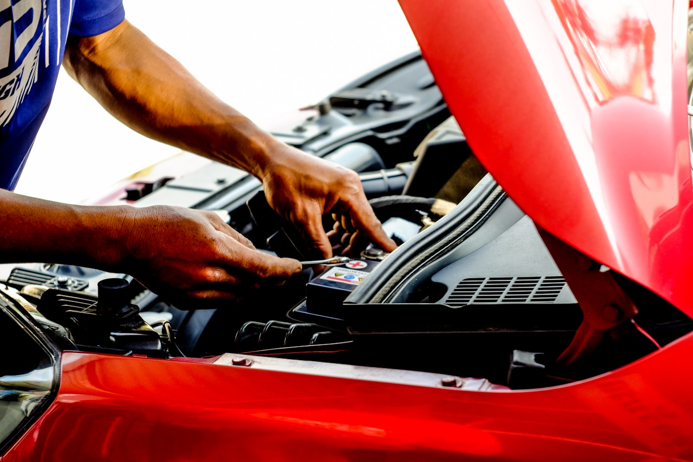 Hands working on a car battery terminal under a raised red hood