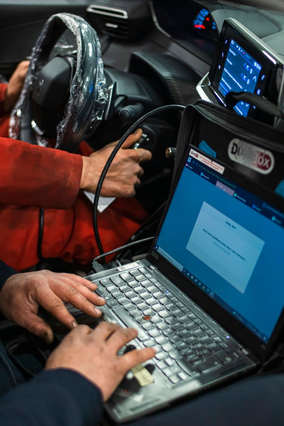 A technician typing on a laptop wired into a car, programming it from the driver's seat
