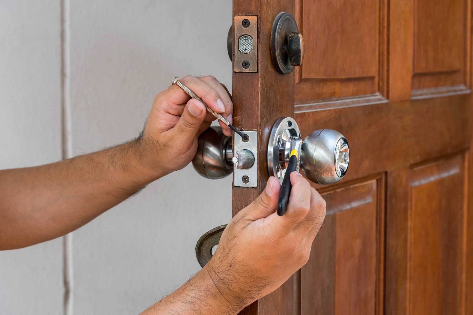 Two hands fitting a chrome knob and latch into a wooden front door with a screwdriver