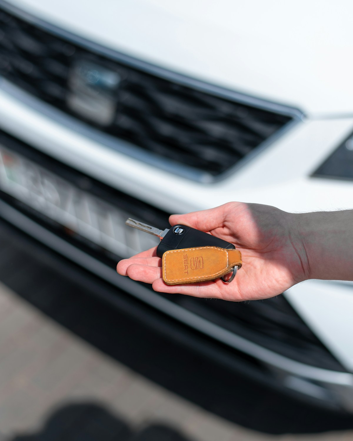 An open hand holding a black car key with a tan leather fob in front of a white car's grille